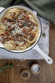 A white cast-iron pan filled with linguine pasta topped with a hearty meat sauce, garnished with fresh herbs and grated cheese. The pan is placed on a rustic wooden surface, and nearby are small wooden bowls containing cracked pepper and coarse salt. A green cloth is partially visible in the background.