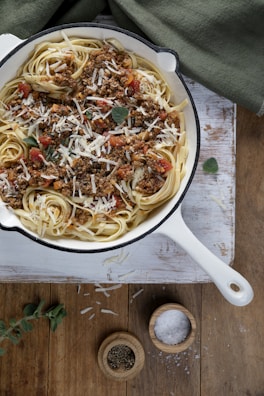 A white cast-iron pan filled with linguine pasta topped with a hearty meat sauce, garnished with fresh herbs and grated cheese. The pan is placed on a rustic wooden surface, and nearby are small wooden bowls containing cracked pepper and coarse salt. A green cloth is partially visible in the background.