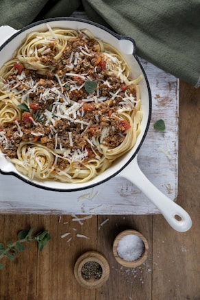 A white cast-iron pan filled with linguine pasta topped with a hearty meat sauce, garnished with fresh herbs and grated cheese. The pan is placed on a rustic wooden surface, and nearby are small wooden bowls containing cracked pepper and coarse salt. A green cloth is partially visible in the background.