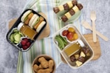 A flat lay image of a lunch spread featuring two bento boxes filled with a variety of foods. The black bento box contains sandwich sliders with lettuce, cucumber slices, a small bottle of juice, and mixed berries. The white bento box includes meat and pineapple skewers, snap peas, grape tomatoes, and another juice bottle. Wooden forks and a bowl of chicken nuggets are also present on a wooden cutting board placed over a striped cloth.