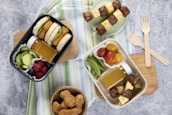 A flat lay image of a lunch spread featuring two bento boxes filled with a variety of foods. The black bento box contains sandwich sliders with lettuce, cucumber slices, a small bottle of juice, and mixed berries. The white bento box includes meat and pineapple skewers, snap peas, grape tomatoes, and another juice bottle. Wooden forks and a bowl of chicken nuggets are also present on a wooden cutting board placed over a striped cloth.