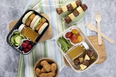 A flat lay image of a lunch spread featuring two bento boxes filled with a variety of foods. The black bento box contains sandwich sliders with lettuce, cucumber slices, a small bottle of juice, and mixed berries. The white bento box includes meat and pineapple skewers, snap peas, grape tomatoes, and another juice bottle. Wooden forks and a bowl of chicken nuggets are also present on a wooden cutting board placed over a striped cloth.