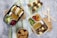A flat lay image of a lunch spread featuring two bento boxes filled with a variety of foods. The black bento box contains sandwich sliders with lettuce, cucumber slices, a small bottle of juice, and mixed berries. The white bento box includes meat and pineapple skewers, snap peas, grape tomatoes, and another juice bottle. Wooden forks and a bowl of chicken nuggets are also present on a wooden cutting board placed over a striped cloth.