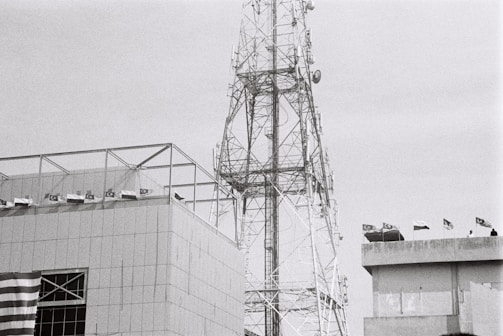 A large telecommunications tower stands next to a modern building. The building has a series of flags on its roof, and part of a patterned flag is visible on the facade. The tower has a complex lattice structure with various antennas attached. The image is in black and white, lending a somewhat industrial and stark appearance.