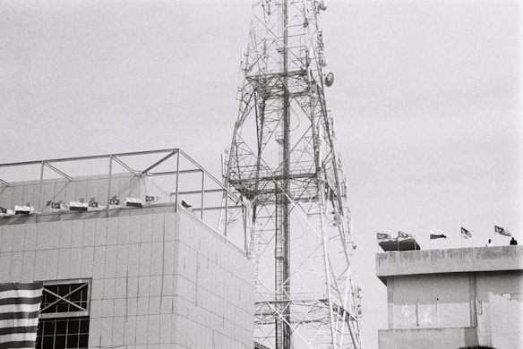 A large telecommunications tower stands next to a modern building. The building has a series of flags on its roof, and part of a patterned flag is visible on the facade. The tower has a complex lattice structure with various antennas attached. The image is in black and white, lending a somewhat industrial and stark appearance.
