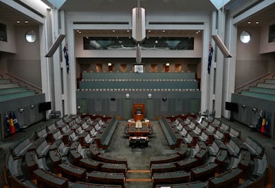 A large, empty parliamentary chamber featuring rows of tiered seating arranged in a semi-circle facing a central speaker's podium. Flags are displayed on both sides of the room, and a digital clock is visible above the speaker's area. The architecture is modern with high ceilings and muted tones.