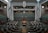 A large, empty parliamentary chamber featuring rows of tiered seating arranged in a semi-circle facing a central speaker's podium. Flags are displayed on both sides of the room, and a digital clock is visible above the speaker's area. The architecture is modern with high ceilings and muted tones.