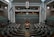A large, empty parliamentary chamber featuring rows of tiered seating arranged in a semi-circle facing a central speaker's podium. Flags are displayed on both sides of the room, and a digital clock is visible above the speaker's area. The architecture is modern with high ceilings and muted tones.