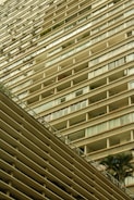 A tall residential building with numerous balconies stretching vertically. The structure features clean lines and a uniform design, with balconies that have glass railings. Some plants are visible on a few balconies, adding a touch of greenery.
