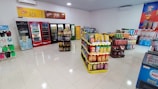 A well-organized convenience store interior featuring several aisles of products. On the left, flip-flops are displayed. The back wall has multiple refrigerators stocked with drinks. In the center, there are shelves filled with snack packages, predominantly chips. To the right, assorted beverages and household items are stacked. The store is brightly lit with a clean, tiled floor.
