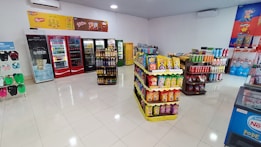 A well-organized convenience store interior featuring several aisles of products. On the left, flip-flops are displayed. The back wall has multiple refrigerators stocked with drinks. In the center, there are shelves filled with snack packages, predominantly chips. To the right, assorted beverages and household items are stacked. The store is brightly lit with a clean, tiled floor.