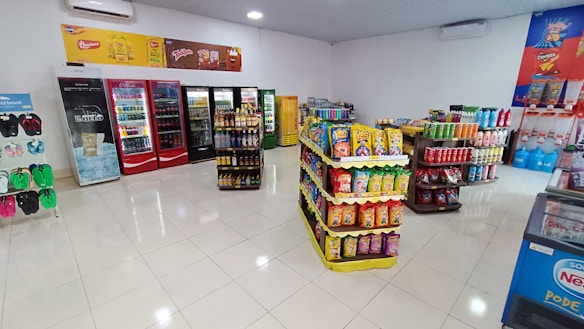 A well-organized convenience store interior featuring several aisles of products. On the left, flip-flops are displayed. The back wall has multiple refrigerators stocked with drinks. In the center, there are shelves filled with snack packages, predominantly chips. To the right, assorted beverages and household items are stacked. The store is brightly lit with a clean, tiled floor.