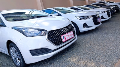 Several white cars are parked in a row on a gravel surface next to a beige wall. The cars appear to be in good condition, and the front car has a noticeable logo and a license plate with 'Patricia' written on it.
