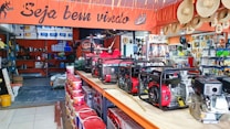A hardware or general store interior featuring several generators displayed prominently on a central counter. Shelves are stocked with various products including paint cans, tools, and other equipment. Straw hats are hung decoratively above the counter. The store is well-lit and organized, with an inviting atmosphere suggested by a 'Seja bem-vindo' sign, which means 'welcome' in Portuguese.