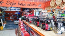 A hardware or general store interior featuring several generators displayed prominently on a central counter. Shelves are stocked with various products including paint cans, tools, and other equipment. Straw hats are hung decoratively above the counter. The store is well-lit and organized, with an inviting atmosphere suggested by a 'Seja bem-vindo' sign, which means 'welcome' in Portuguese.