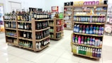 A grocery store interior featuring shelves filled with various products including bottles of alcoholic beverages and cleaning supplies neatly arranged. The floor is tiled, and there is a soft, well-lit ambiance.