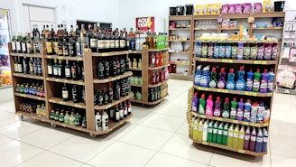 A grocery store interior featuring shelves filled with various products including bottles of alcoholic beverages and cleaning supplies neatly arranged. The floor is tiled, and there is a soft, well-lit ambiance.
