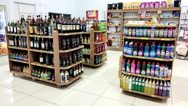 A grocery store interior featuring shelves filled with various products including bottles of alcoholic beverages and cleaning supplies neatly arranged. The floor is tiled, and there is a soft, well-lit ambiance.