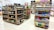 A grocery store interior featuring shelves filled with various products including bottles of alcoholic beverages and cleaning supplies neatly arranged. The floor is tiled, and there is a soft, well-lit ambiance.