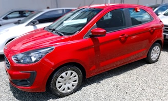 A red compact car is parked on a gravel surface. It features a modern design with smooth lines and a shiny finish. The vehicle is surrounded by a few other cars, indicating a parking lot setting. The side view mirrors and windows are present, and the car appears to be in good condition.