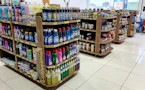 Several shelves are filled with various cleaning products, snacks, and household items. The floor is tiled, and there is ample lighting coming through the store. Each shelf is well-organized with items neatly stacked and price tags visible.