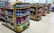 Several shelves are filled with various cleaning products, snacks, and household items. The floor is tiled, and there is ample lighting coming through the store. Each shelf is well-organized with items neatly stacked and price tags visible.
