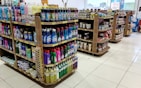Several shelves are filled with various cleaning products, snacks, and household items. The floor is tiled, and there is ample lighting coming through the store. Each shelf is well-organized with items neatly stacked and price tags visible.