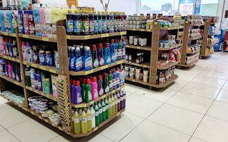Several shelves are filled with various cleaning products, snacks, and household items. The floor is tiled, and there is ample lighting coming through the store. Each shelf is well-organized with items neatly stacked and price tags visible.