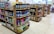 Several shelves are filled with various cleaning products, snacks, and household items. The floor is tiled, and there is ample lighting coming through the store. Each shelf is well-organized with items neatly stacked and price tags visible.