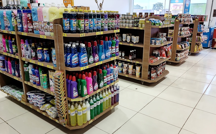 Several shelves are filled with various cleaning products, snacks, and household items. The floor is tiled, and there is ample lighting coming through the store. Each shelf is well-organized with items neatly stacked and price tags visible.