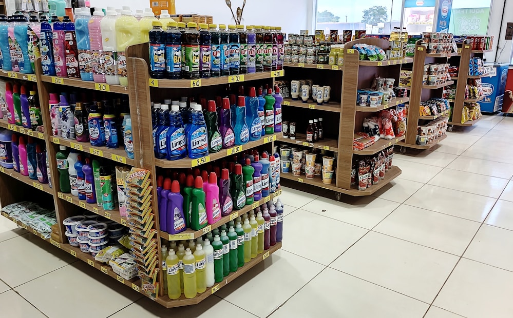 Several shelves are filled with various cleaning products, snacks, and household items. The floor is tiled, and there is ample lighting coming through the store. Each shelf is well-organized with items neatly stacked and price tags visible.