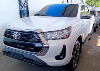 A white pickup truck is parked indoors. The vehicle has a shiny chrome grille and prominent brand emblem at the front. Its polished surface reflects the surroundings, indicating it's well-maintained. The background reveals part of another vehicle and a blue cleaning product on a shelf.