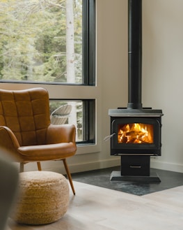 A cozy corner of a room featuring a modern wood-burning stove with a visible fire, a large window providing a view of trees outside, a comfortable brown leather chair, and a woven pouf on a light wooden floor.