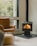 A cozy corner of a room featuring a modern wood-burning stove with a visible fire, a large window providing a view of trees outside, a comfortable brown leather chair, and a woven pouf on a light wooden floor.