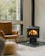 A cozy corner of a room featuring a modern wood-burning stove with a visible fire, a large window providing a view of trees outside, a comfortable brown leather chair, and a woven pouf on a light wooden floor.