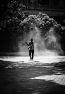 A person is sweeping an outdoor paved area, surrounded by dust and debris creating a hazy atmosphere. The scene is set in black and white, with shadows from trees partially covering the ground and foliage in the background.