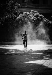A person is sweeping an outdoor paved area, surrounded by dust and debris creating a hazy atmosphere. The scene is set in black and white, with shadows from trees partially covering the ground and foliage in the background.