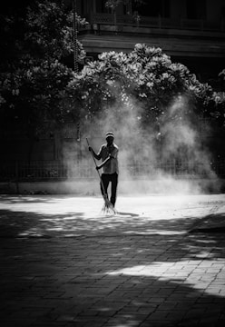 A person is sweeping an outdoor paved area, surrounded by dust and debris creating a hazy atmosphere. The scene is set in black and white, with shadows from trees partially covering the ground and foliage in the background.
