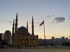 A large mosque with multiple minarets and a prominent blue dome is pictured in an urban setting at sunset. The structure is silhouetted against a soft, light blue and orange sky. Next to it, a Lebanese flag waves on a tall flagpole. Surrounding buildings and a few cars on the street are visible, completing the cityscape.