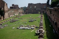 A historical ruin with ancient brick walls surrounding a rectangular open space. The area is grass-covered, and scattered with numerous stone artifacts and remnants of past structures. The walls are partially intact, with some arches and windows still visible, indicating architectural details.