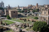 A panoramic view of ancient Roman ruins with several historical buildings featuring classical architecture. The scene includes structures with columns, arches, and domes, surrounded by green spaces and trees. In the distance, more historical buildings and the urban landscape of the city can be seen.
