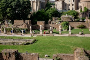 A historical site with ancient Roman statues lined up along a pathway. The background features old stone ruins and lush green trees and bushes. Visitors are walking along the path, some admiring the sculptures.