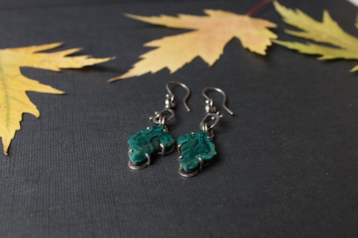 A pair of silver earrings with green, irregularly shaped stones placed on a dark textured surface. Surrounding the earrings are a few autumn leaves in shades of yellow and pale orange.