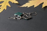 A pair of silver metal earrings with green stones placed on a textured dark surface. There are two dried yellow maple leaves in the corners, adding a natural touch to the composition.