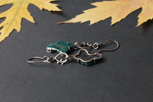 A pair of silver metal earrings with green stones placed on a textured dark surface. There are two dried yellow maple leaves in the corners, adding a natural touch to the composition.