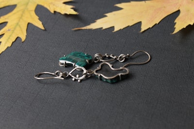 A pair of silver metal earrings with green stones placed on a textured dark surface. There are two dried yellow maple leaves in the corners, adding a natural touch to the composition.
