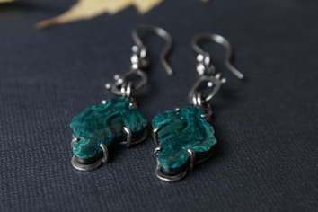 A pair of elegant earrings featuring asymmetrical, green crystal stones set in metal hooks. The stones showcase swirling patterns in shades of green, adding texture and depth. The dark background highlights the earrings, enhancing their intricate details.