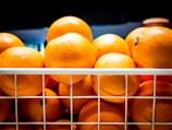 A number of fresh, ripe oranges stacked together inside a white wire basket. The oranges appear to be brightly colored and are placed under a light, which highlights their vibrant skin texture.