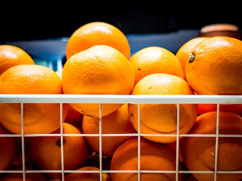 A number of fresh, ripe oranges stacked together inside a white wire basket. The oranges appear to be brightly colored and are placed under a light, which highlights their vibrant skin texture.