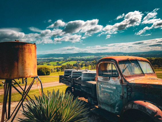 A sturdy water truck parked in front of a sunlit rural landscape with fields and farm buildings.